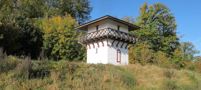Foto: rekonstruierter Limes-Wachturm mit Brüstung und weiß-rotem Mauerstrich hinter grasbewachsenem Wall („von Germanien aus”).