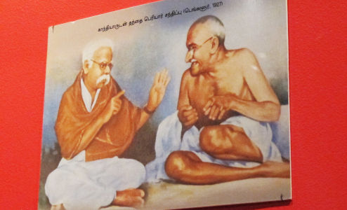 Ramasami und Gandhi als bereits etwas ältere Männer auf einem nachkoloriert und retouschiert wirkenden Foto. Ramasami erklärt gerade etwas.