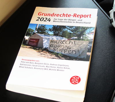 Ein Buch liegt auf einem Tischchen eines Regionalzugs: „Grundrechte-Report“ in Rot, „2024“ in Schwarz, auf weißem Grund mit einem Foto einer Aktion zur Verteidigung des Asylrechts.