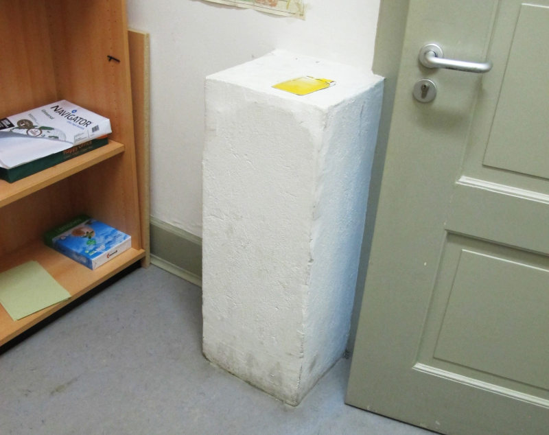 Rechts eine fast geschlossene Zimmertür, daneben ein etwa ein Meter hoher, weiß getünchter Betonblock mit einem Querschnitt von vielleicht 30×30 cm, wieder daneben ein Regal mit ein paar Packen Kopierpapier.