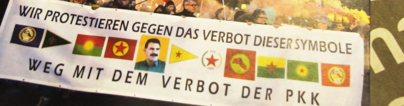 Foto: "Wir protestieren gegen das Verbot dieser Symbole" über einem Satz von Fahnen und Wimpeln kurdischer Organisationen und deinem Öcalan-Portrait.