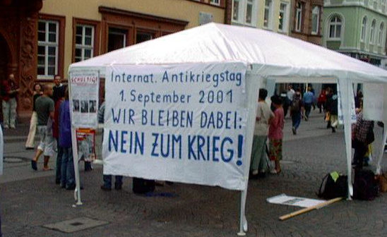 Ein Gazebo mit einem Transparent dran: „Internationaler Antikriegstag 1. September 2001.  Wir bleiben dabei: Nein zum Krieg“, dahinter eine Fußgängerzonenszene.