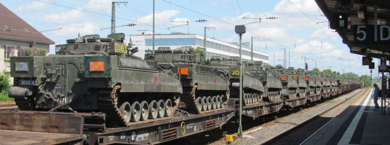 Rechts im Bild ein Bahnsteig, links ein endloser Güterzug.  Auf jedem Wagen stehen zwei Panzer.