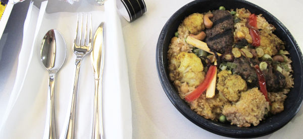 A plastic bowl with rice, cauliflower, chickpea, kofta and a few other things in it.  Next to it, a selection of plastic cutlery.