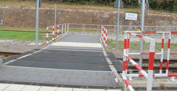 A paved crosswalk over a railway track with some red-and-white warning fencing around it.  There is a plain white sign saying: “Actung! Anfahrender Zug“
