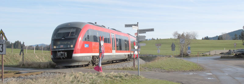 A small red train in a lush, green landscape at a tiny railway crossing with a minuscle signpost.