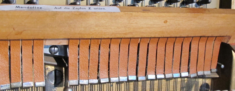 Foto von ca. 20 Lederbändern mit aufgepressten Metallplättchen über Klaviersaiten mit einem Holzbügel mit der Aufschrift „Mandoline auf die Zapfen Z setzen”.