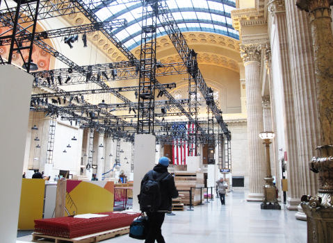 A large hall with faux columns with Corinthian capitals and other historicising decor.  Two people cross it, various construction material takes up most of the floor.