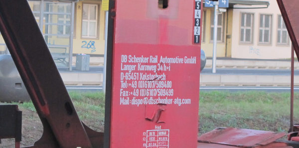 Eine rostfarbene Strebe eines Güterwagens mit aufgesprühten weißen Buchstaben „DB Schenker Rail Automotive GmbH [...] Mail: dispo@dbschenker-atg.com“