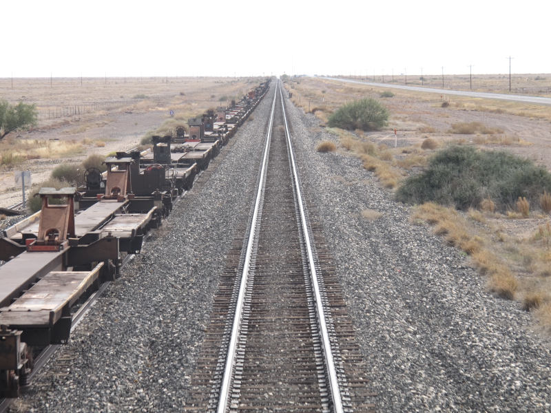 A rail goes straight to the horizon. Right of it is another rail with a long string of flatbed cars on it.