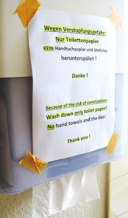 Ein Papierhandttuchspender mit einer aufgeklebten zweisprachigen Nachricht: „Wegen Verstopfungsgefahr: Nur Toilettenpapier KEIN Handtuchpapier und ähnliches herunterspülen! Danke!