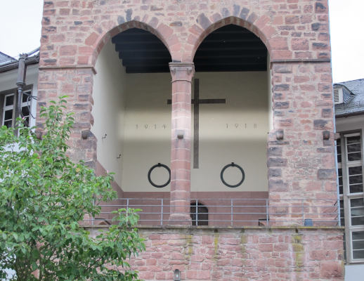 Ausschnitt aus einen grob gemauerten Turm.  Es öffnet sich eine Art Loggia, an deren weiß verputzer Rückwand die Zahlen 1914 und 1918 rechts und links von einem Kreuz zu erkennen sind.