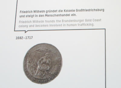 Screenshot mit Siegel und zweisprachigem Text: „Friedrich Wilhelm gründet die Kolonie Großfriedrichsburg und steigt in den Menschenhandel ein, 1682-1712.