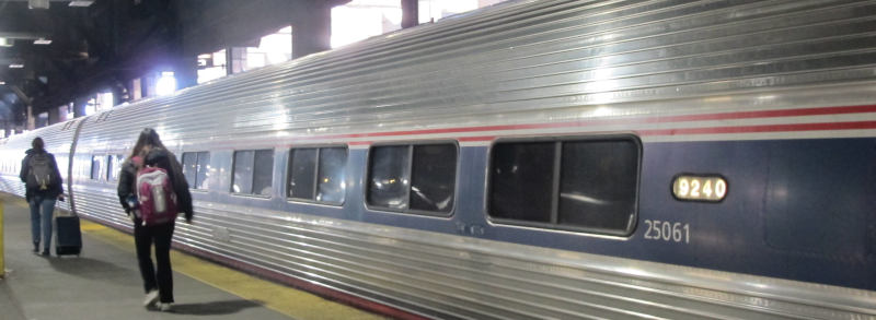 A few cars of a train in corrugated-alumnium look with a blue stripe connecting the windows.