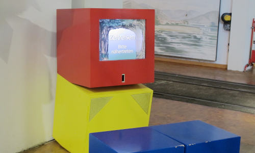 Vier Klötze in kräftigen Farben, Sitzklötze in Blau, ein Monitorklotz in rot.  Auf dem Monitor eine Anzeige „Zeitreisen/Bitte nähertreten“.