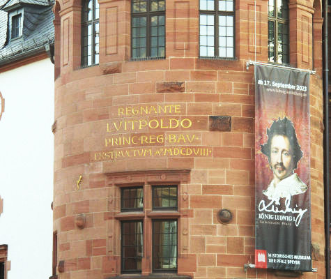 Teil-Außenansicht des historischen Museums; auf einer Sandsteinmauer hängt eine Werbefahne für die Ludwig-Ausstellung, links daneben in Gold in die Wand eingelegt: „Regnante Luitpoldo Princ Reg Bav Exstructum AºMDCDVIII”