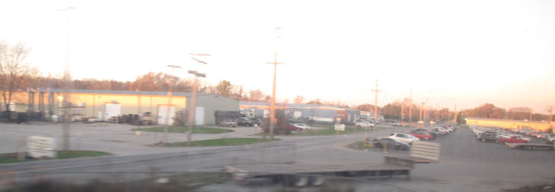 A straight road with utility poles accompanying it, a parking lot, and several halls made of corrugated sheet, there's some amount of motion blur.