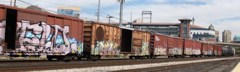 A freight train make up primarily of boxcars in front of a few urban houses.  The door of the second boxcar visible is open.
