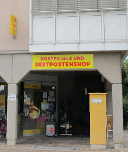An einer Arkade eines 70er-Jahre-Baus hängt ein Schild „Postfiliale und Restpostenshop“, davor ein Gelber Postkasten.
