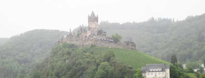 Eine Burg mit vielen Türmchen auf einem mit Wein und Gestrüpp bewachsenen Hügeln vor im Hintergrund blasseren Hügeln.