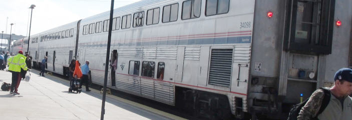 A train with four doubledecker cars, corrugated sheet look and a red-and-blue decorative strip on the sides.  At the right, the rear rend of the train faces the viewer with two red lights.