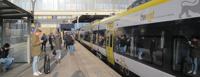 Foto: Ein Zug steht mit geschlossenen Türen auf dem Bahnsteig, etliche Menschen in verschiedenen Stadien der Verwirrung davor.