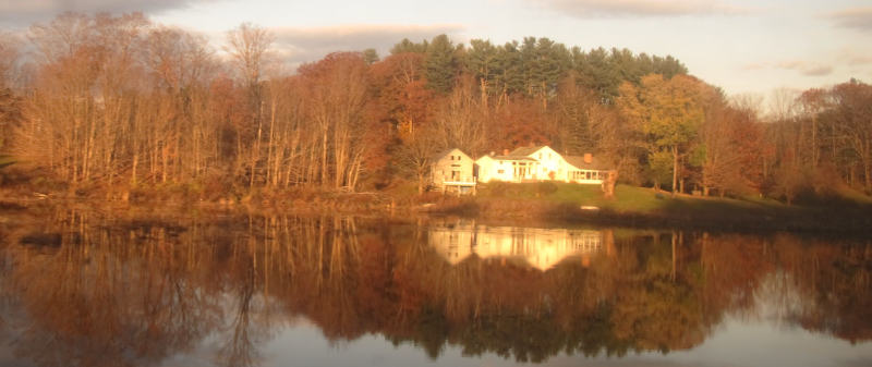 A several-bedroom house standing alone in front of a small grove and behind a lake which reflects the scene.  Everything is bathed in warm light.