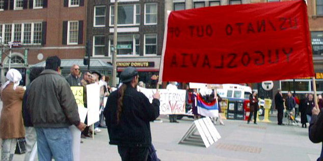 Eine Kundgebung mit einer guten Handvoll Menschen vor Stadthäusern.  Ein rotes Transparent, auf dem „US/NATO out of Yugoslavia“ steht, ist von hinten zu sehen.