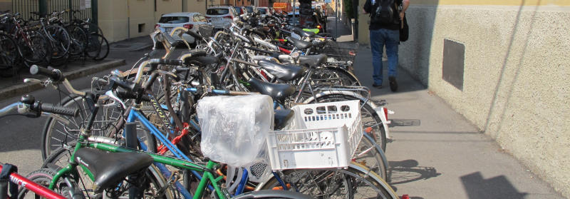 ca. 20 Fahrräder stehen nebeneinander geparkt an einer Straße.