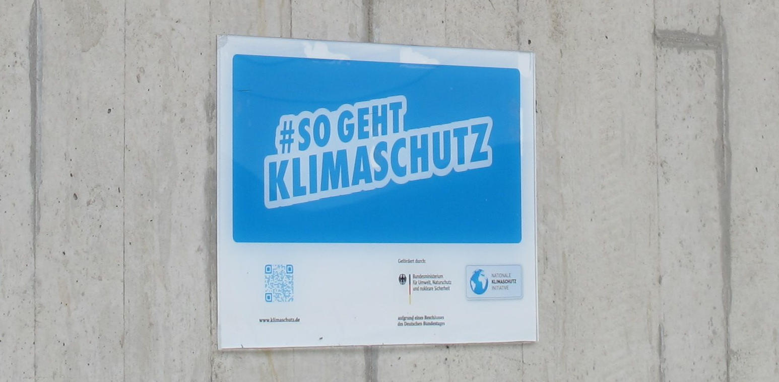 Ein Schild an einer Betonwand, groß #SO GEHT KLIMASCHUTZ, dann mit Logos des Umweltministeriums und der Nationalen Klimaschutz-Inititative sowie einem QR-Code und dem kleinen Schriftzug www.klimaschutz.de.