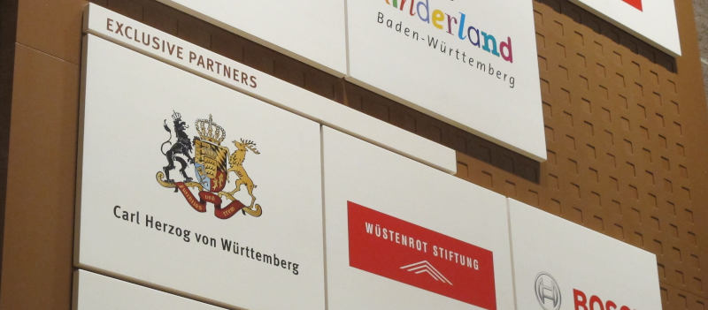 Auf einer braunen Wand angebrachte weiße Schilder mit diversen Logos unter der Überschrift „Exclusive Partners“ („Wüstenrot-Stiftung“, „Bosch“).  Ganz vorne jedoch ein Wappen mit Krönchen und allem, darunter „Carl Herzog von Württemberg“.