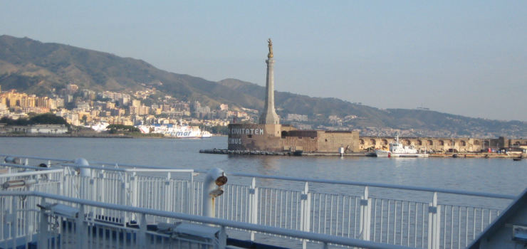 A shore covered with houses, the sea in front of it.  From the right, a pier enters the image, with a pillar at its end.  In the foreground, a ship's railing is visible.