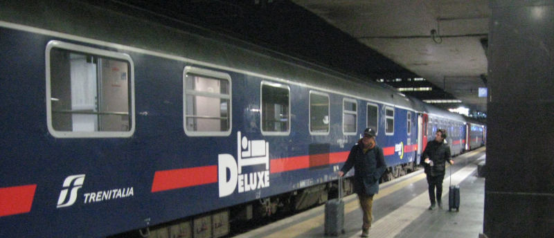 A blue sleeper car marked “deluxe” on a platform at night.  Two persons with trolleys walk alongside, clearly looking forward to finding their rolling beds.