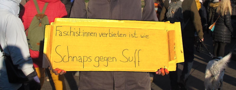 Eine Person hält vor dem Hintergrund einer Demo eine Pappe: „Faschist:innen verbieten ist wie Schnaps gegen Suff“.