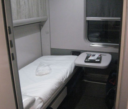 A view into a sleeper cabin through its door; a single bunk with white linen.