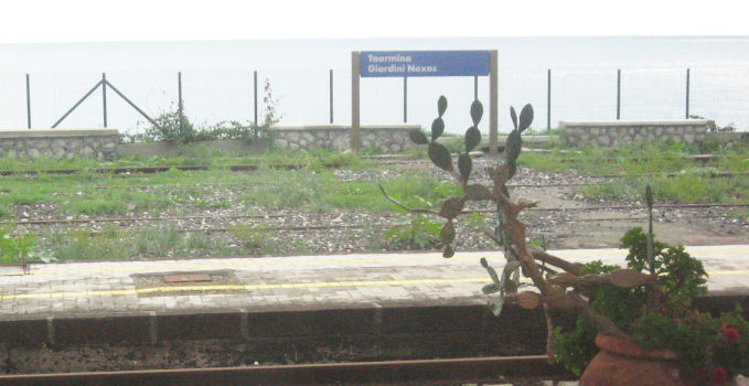 A platform and a sign “Taormina Giadini Nexos“ in front of a grayish ocean.  A potted cactus grows into the image.