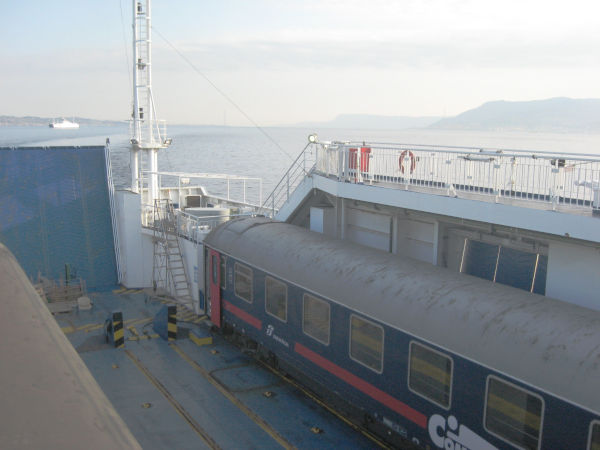 View into a ferry with rails and a sleeper car.  Behind that, the ocean and the landscape of the strait of Messina.