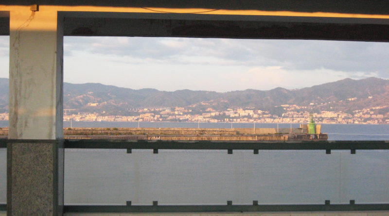 A hilly landscape with many houses beyond a piece of blue sea, all viewed through a concrete construction belonging to a railway station.