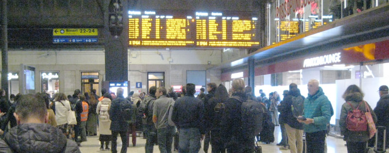 In a staion hall: A large dot-matrix display showing train connections, and lots of people staring at it, seen from behind.