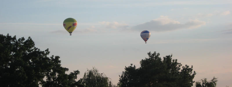 Zwei bunte Heißluftballons am Abendhimmel; den unteren Bildrand bilden Bäume im Schattenriss