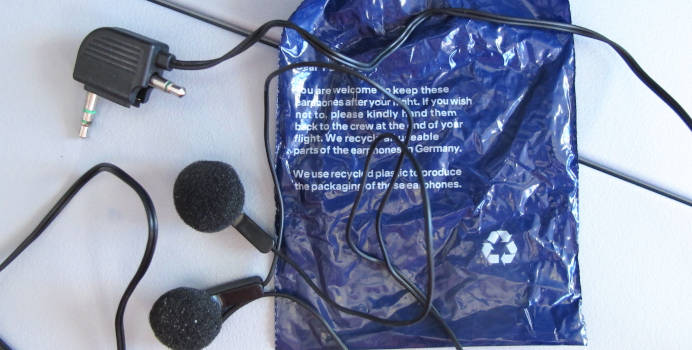 Ein in-ear-Kopfhörer mit einem Klinkenstecker, der durch Klappen in einen der alten zweisteckrigen Flugzeughörer verwandelt werden kann.  Darunter ein leerer tiefblauer Plastikbeutel mit weißem Text und Recycling-Logo.
