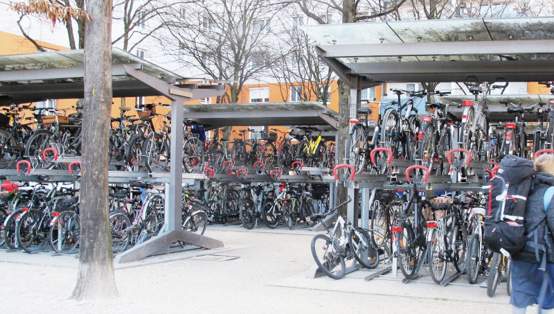 Vier Stahlkonstruktionen mit nach innen geneigten Plexiglasdächern. Seitlich offen, stehen in zwei Stockwerken auf beiden Seiten Fahrräder, vielleicht insgesamt 80 pro Ständer. Praktisch alle Plätze sind belegt.