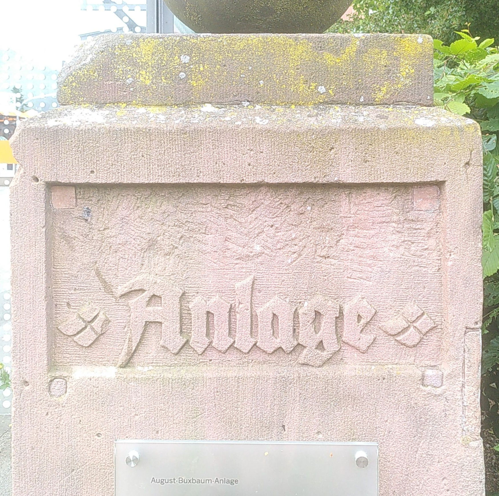Ein Sandsteinpfeiler mit einem zweizeiligen Textfeld.  Die erste Zeile ist ausgemeißelt, die zweite zeigt in Fraktur zentriert „Anlage“.