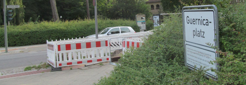 Ein rot-weißer Baustellenzahn, ein Stück daneben ein weißes Schild „Guernica-Platz“.
