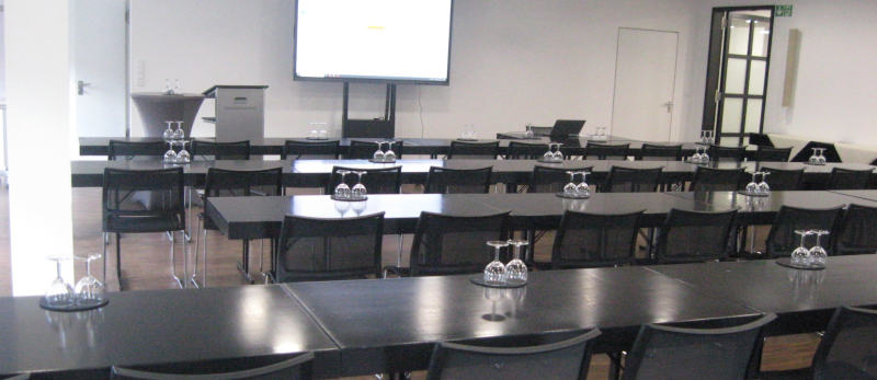 Ein Besprechungsraum mit einem leeren Monitor an der breiten Strirnseite.  Auf den schwarzen, glänzenden Tischen stehen umgedrehte Weingläser.